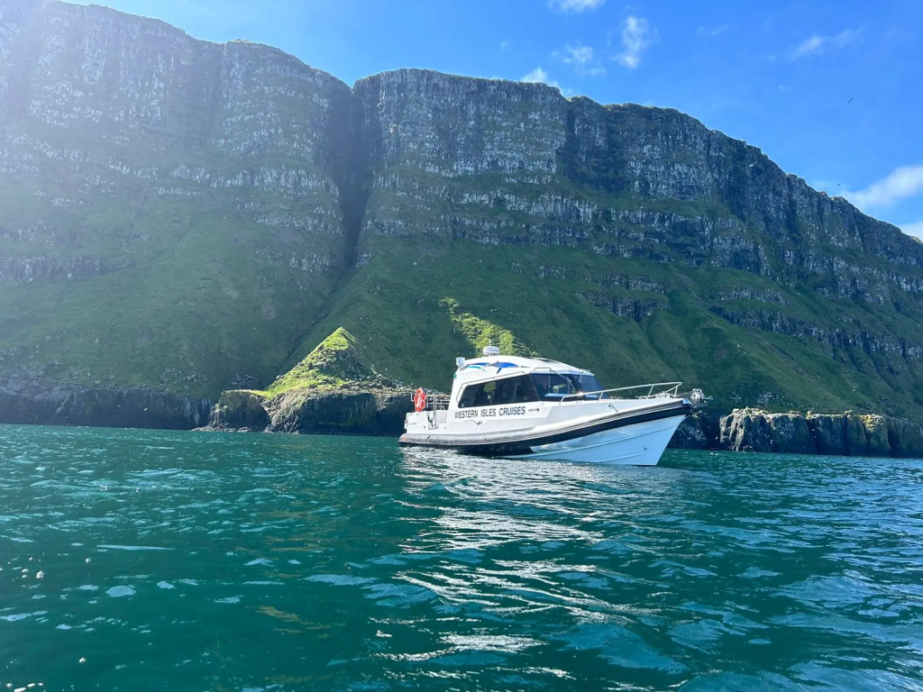 White boat on blue water near a rocky, green mountain under a clear blue sky.