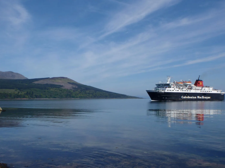 imgi_1_calmac-1 Ferry on calm water with a hilly landscape and blue sky in the background.