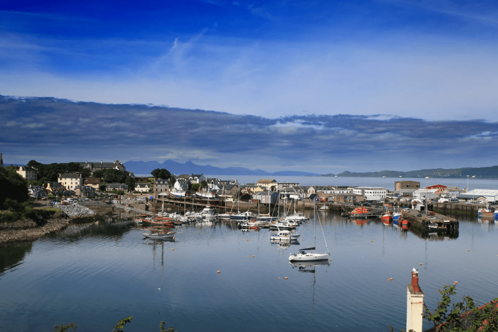 Scenic view of a harbor with boats, buildings, and distant mountains under a blue sky.