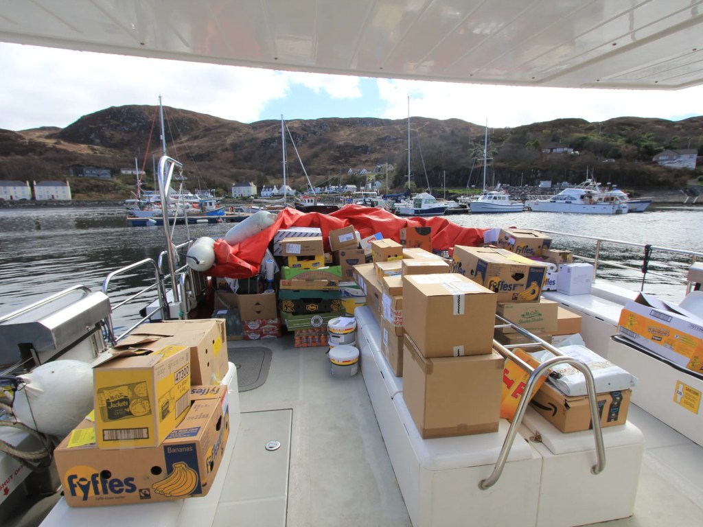 Boat deck loaded with cardboard boxes and supplies, docked near a small harbor.