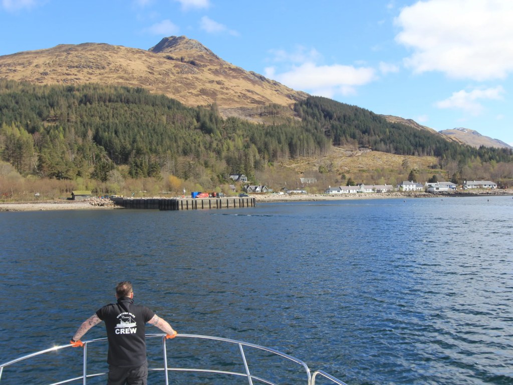 Person on boat looking at mountainous coastline and pier under a blue sky.