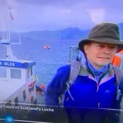 Person in outdoor gear on boat in a loch, cloudy sky.