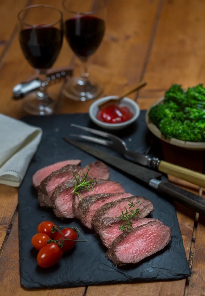 Sliced steak with tomatoes, broccoli, and red wine on a wooden table.