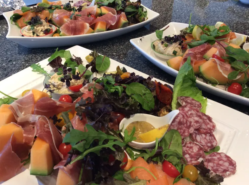 Platters with assorted charcuterie, melon, prosciutto, greens, and dips on a granite counter.