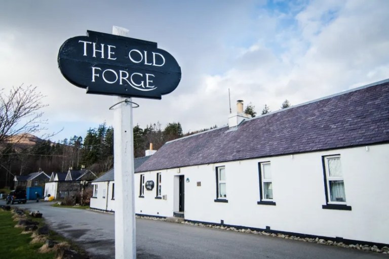 Sign for 'The Old Forge' beside a white building under a cloudy sky.