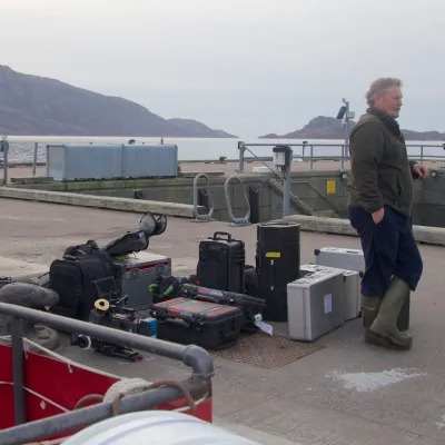 Person stands on a dock with equipment cases; mountains and water in background.