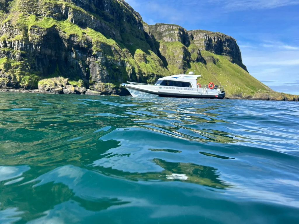 Boat cruising near rocky, grassy cliffs on a sunny day.