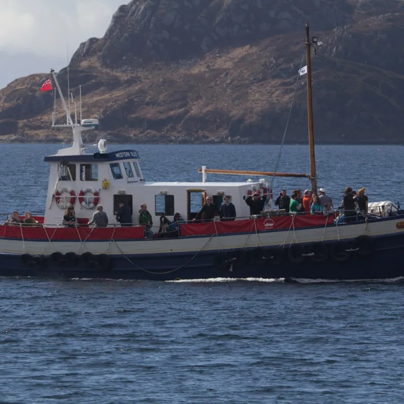 Boat with passengers on water, rocky hill in background.
