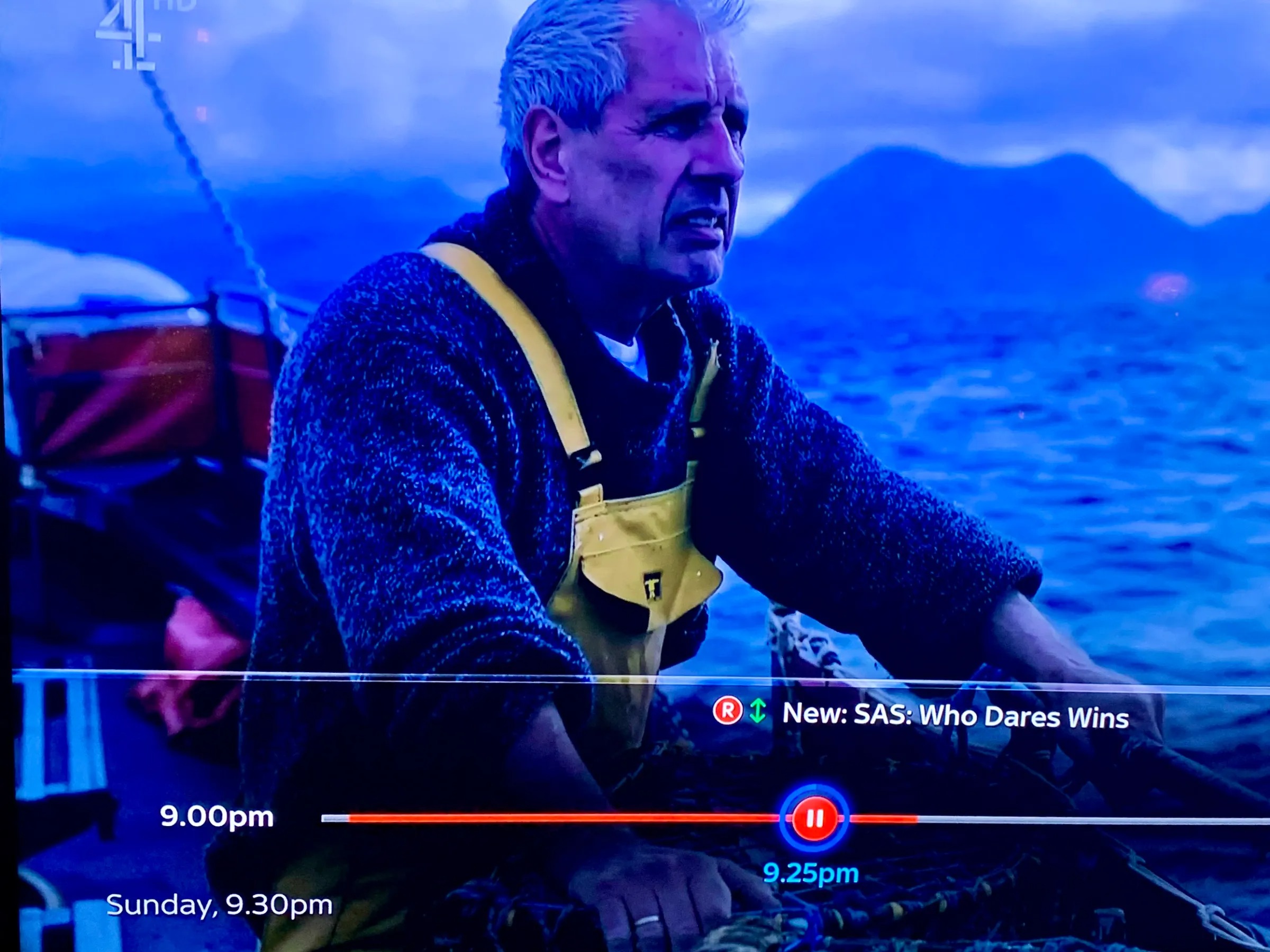 Man in yellow overalls on a boat at sea with mountains in background.
