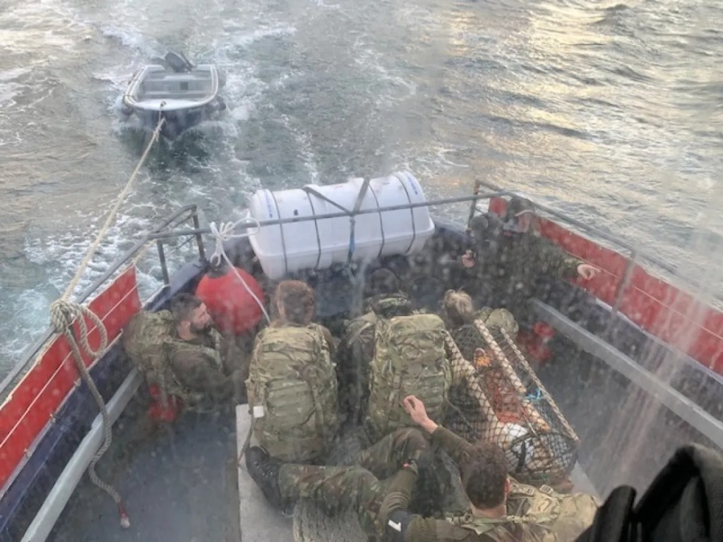 People in military attire on a boat towing a small dinghy on water.