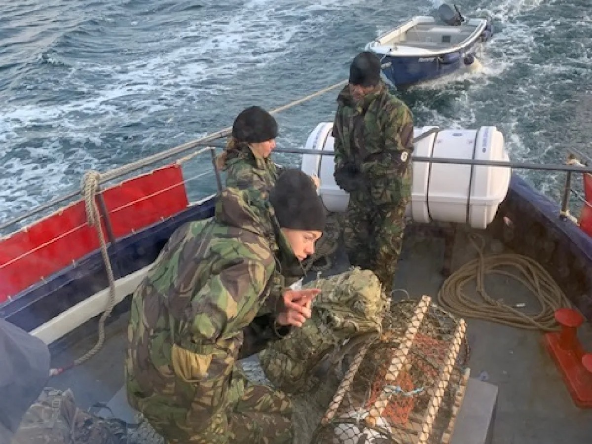 People in camouflage gear on a boat handling a net with a smaller boat nearby.