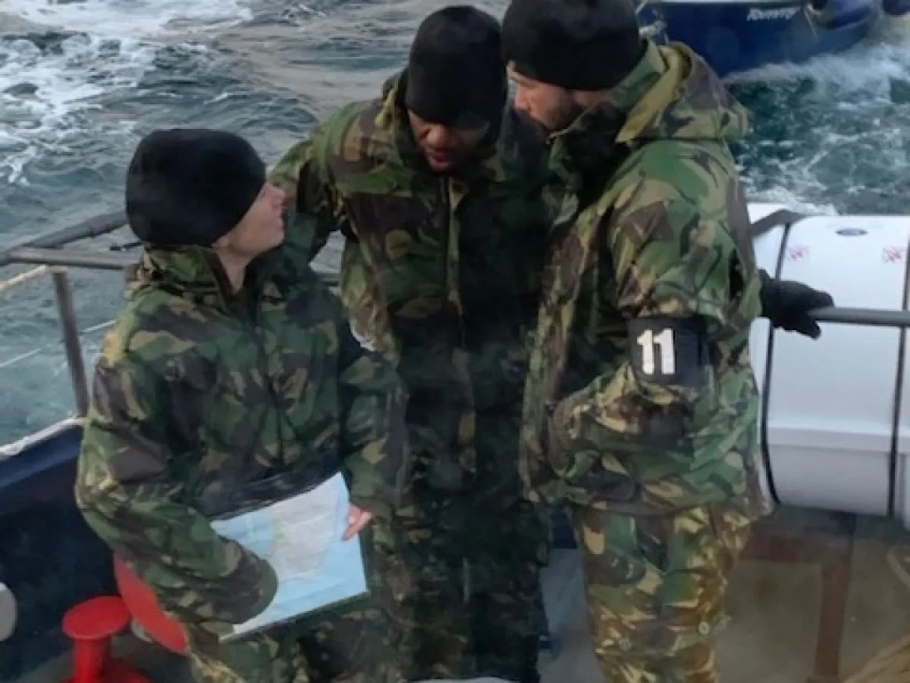 Three people in camouflage gear on a boat, holding a map and talking.