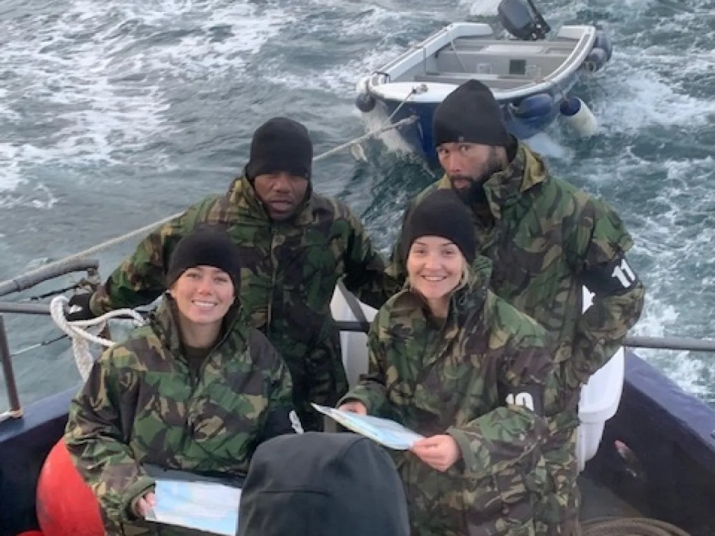 Four people in camo gear on a boat holding maps over the sea.