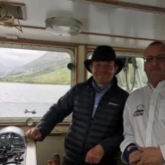Two men on a boat with scenic lake and hills visible through windows.