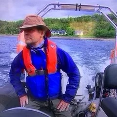 Person with hat and life jacket steering a boat on a lake.
