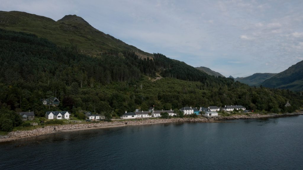 Houses along a forested hillside by a calm lake, with mountains in the background.