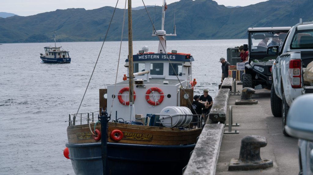 Boat docked at pier with people and vehicle, mountains in background.