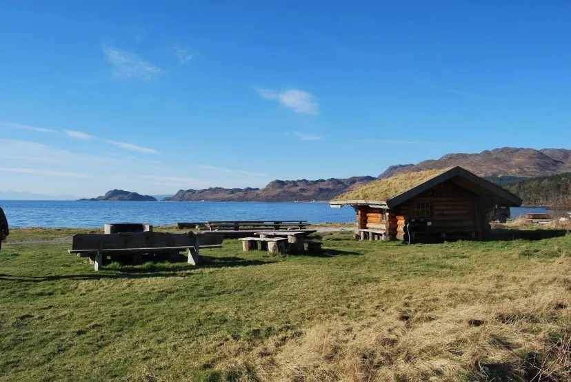 Lakeside cabin with picnic tables, grassy area, and distant mountains under clear blue sky.