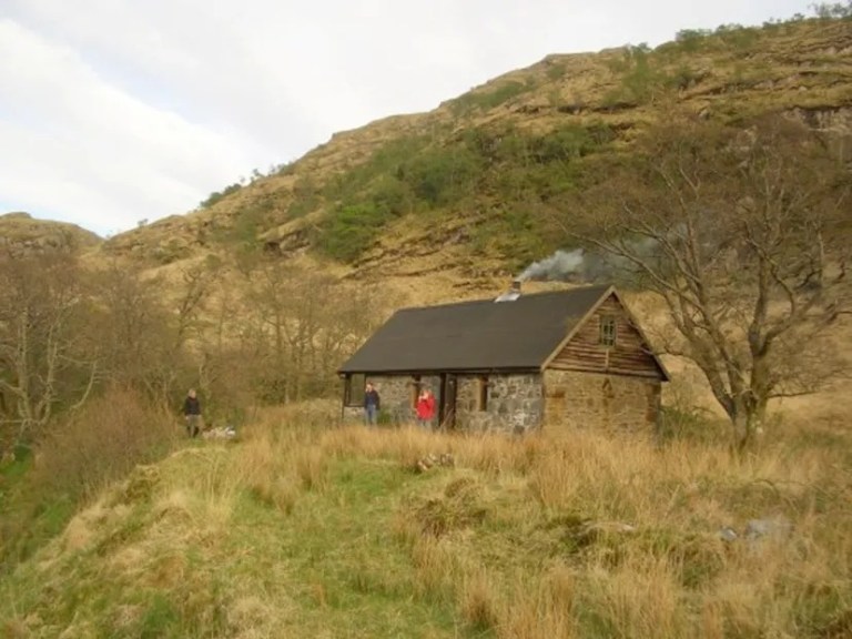 Stone cabin with smoke in a hilly landscape, people nearby.