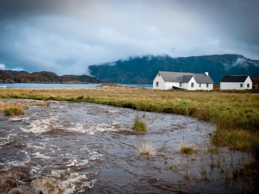 Riverside landscape with two white houses, grassy field, and mountains in the background under cloudy sky.