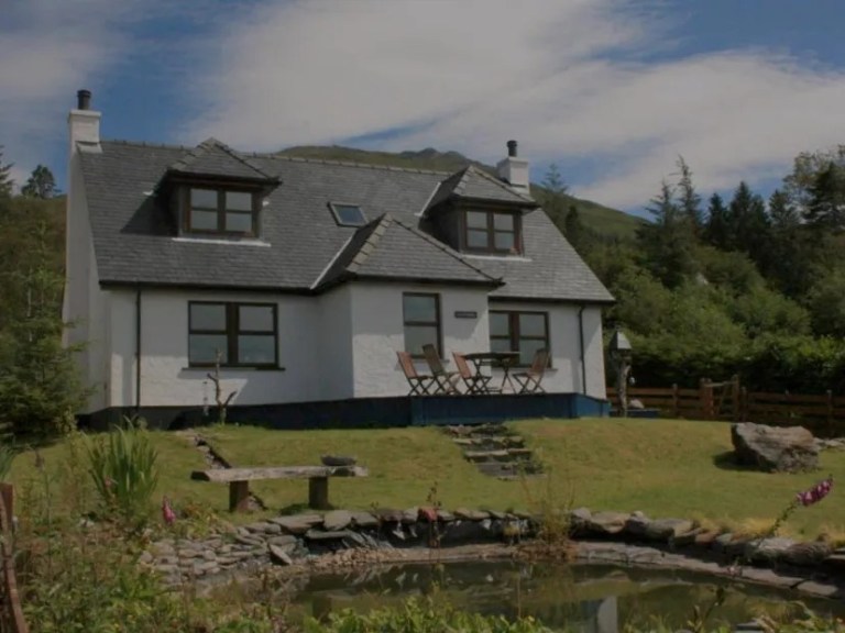 Charming white cottage with attic windows, surrounded by trees and a small pond in front.