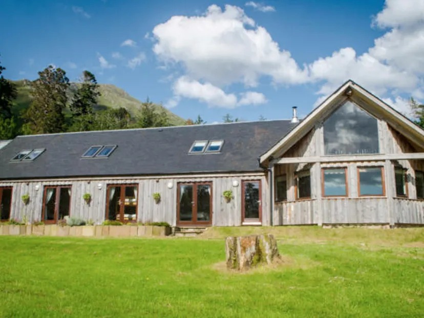 Wooden house with large windows and sloped roof, set in a grassy area with mountains in the background.