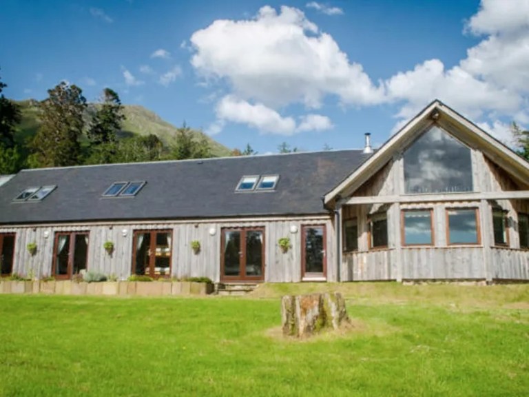 Wooden house with large windows and sloped roof, set in a grassy area with mountains in the background.