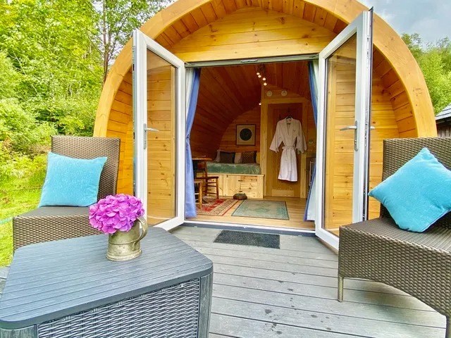 Cozy wooden cabin with open double doors, wicker chairs, and table with pink flowers.