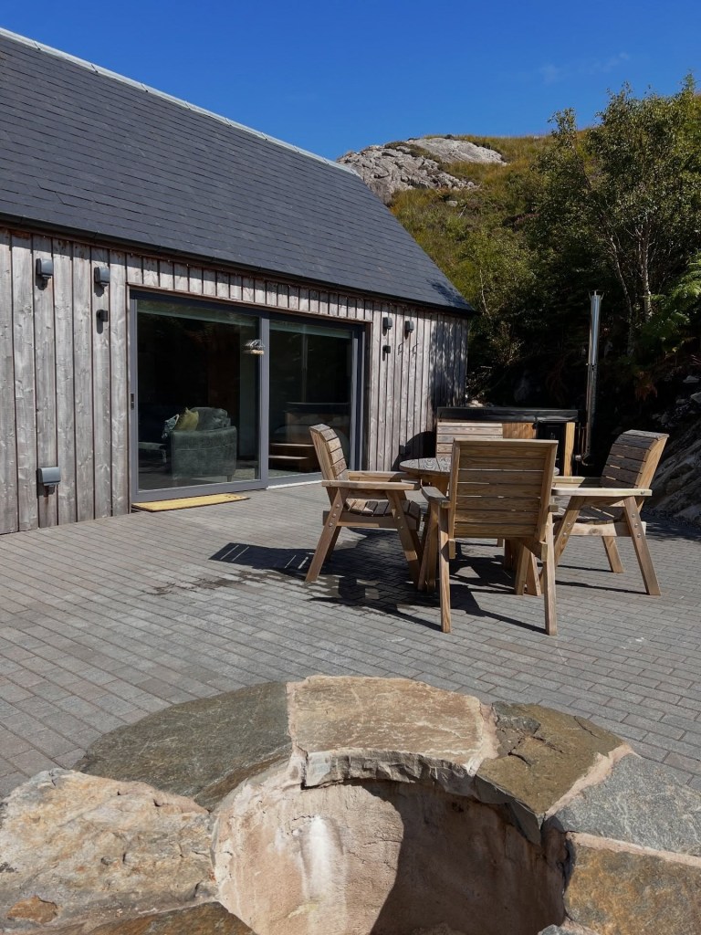Wooden cabin with patio, outdoor table, chairs, and stone fire pit under blue sky.