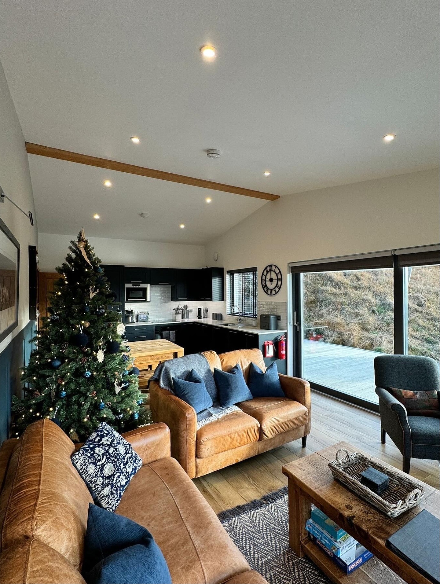 Modern living room with Christmas tree, brown sofas, and view of kitchen and outdoor area.