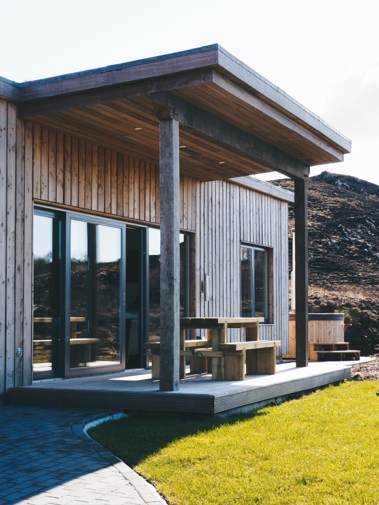 Modern wooden cabin with a porch, large glass doors, and outdoor seating on a sunny day.