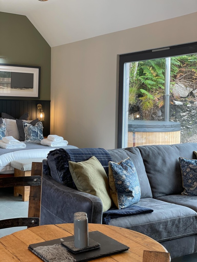 Cozy room with bed, sofa, and pillows near large window showing greenery outside.