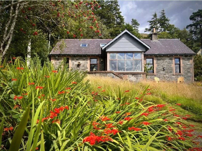 Stone cottage with large window, surrounded by trees and red flowers in foreground.